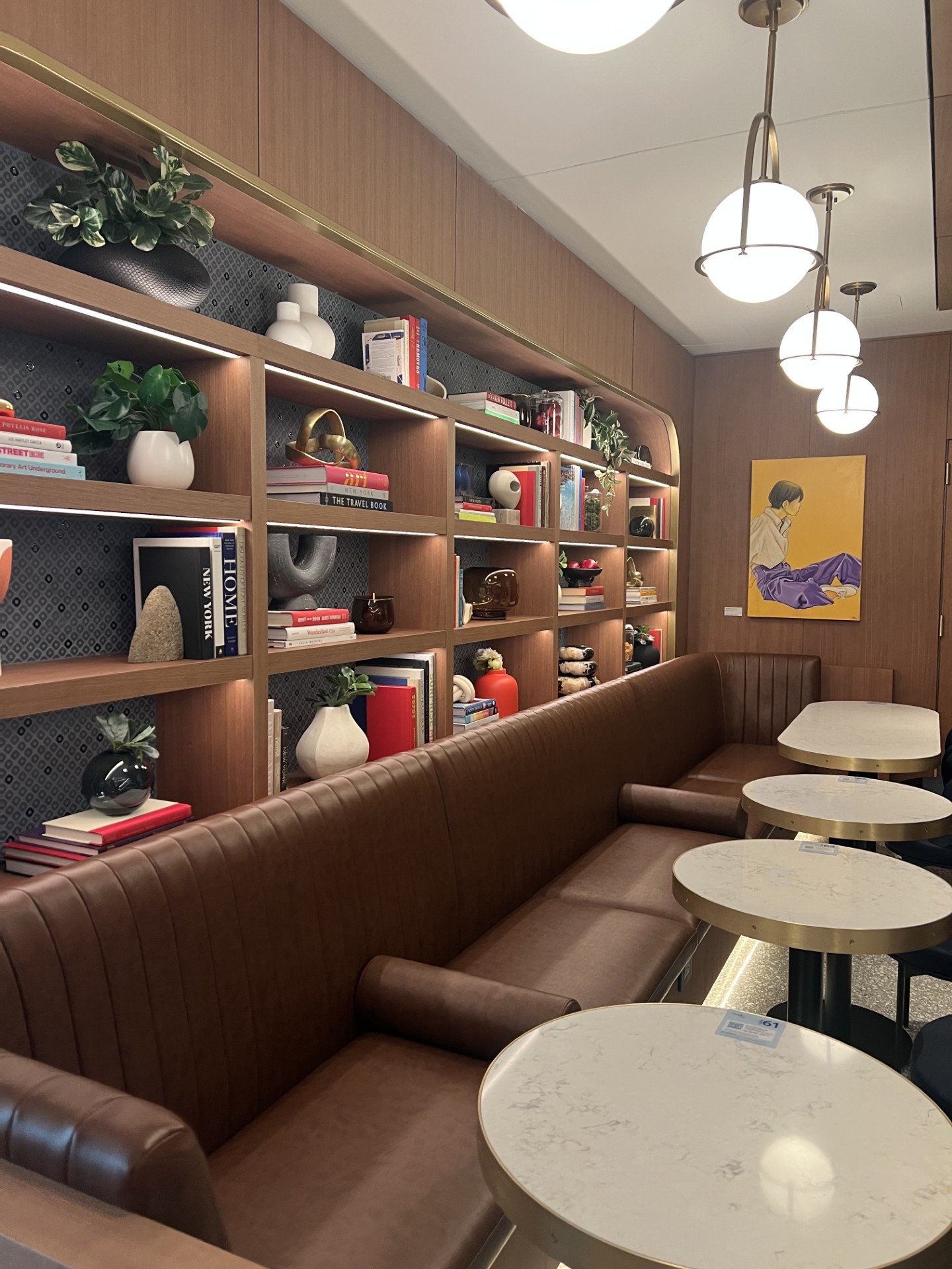 A seating area in the new JFK Capital One airport lounge has cafe-style seating with a banquette and several round tables, with shelving nooks filled with tasteful objects and art.